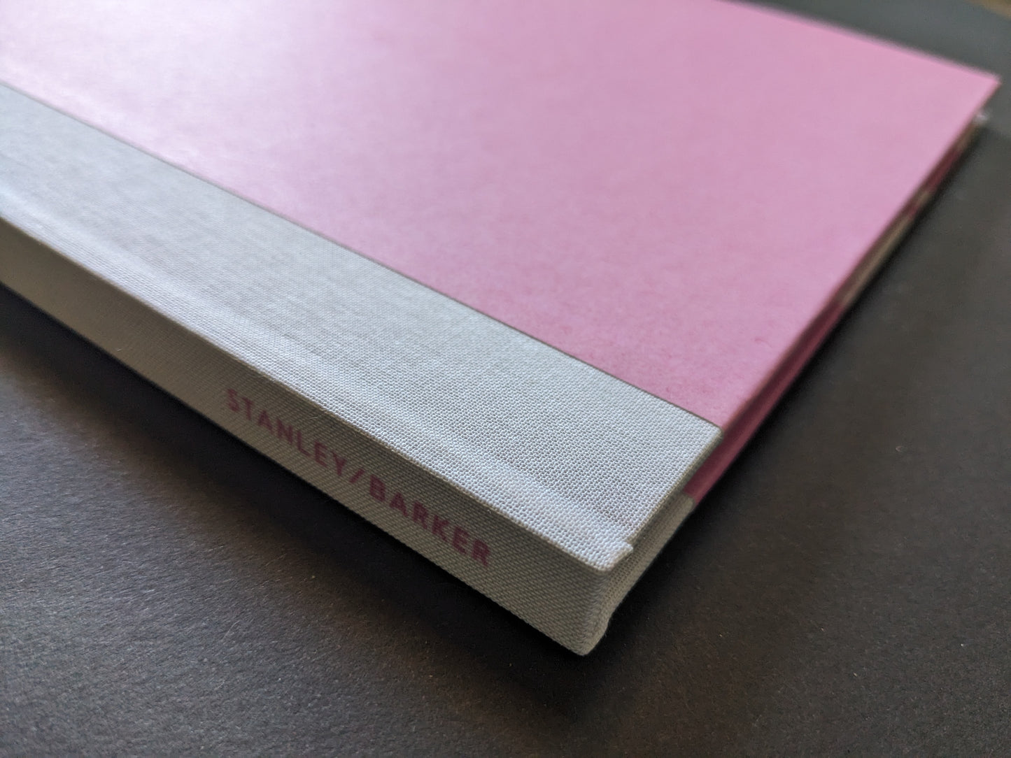 Cloth-bound spine of "Flowers Drink the River" with Stanley/Barker logo in pink on grey fabric.