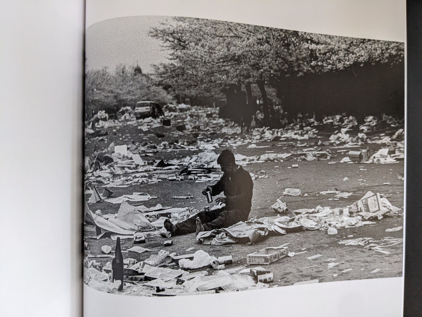 A solitary figure sits on the ground amid a landscape strewn with trash and debris, conveying isolation and desolation.