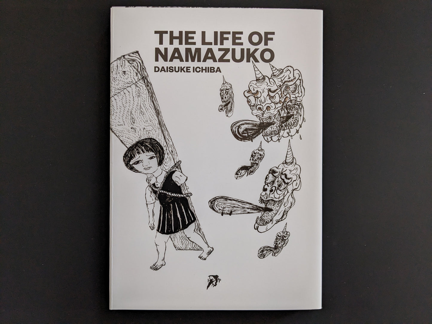 Front cover of The Life of Namazuko by Daisuke Ichiba, showing a girl carrying a plank surrounded by surreal demon heads.