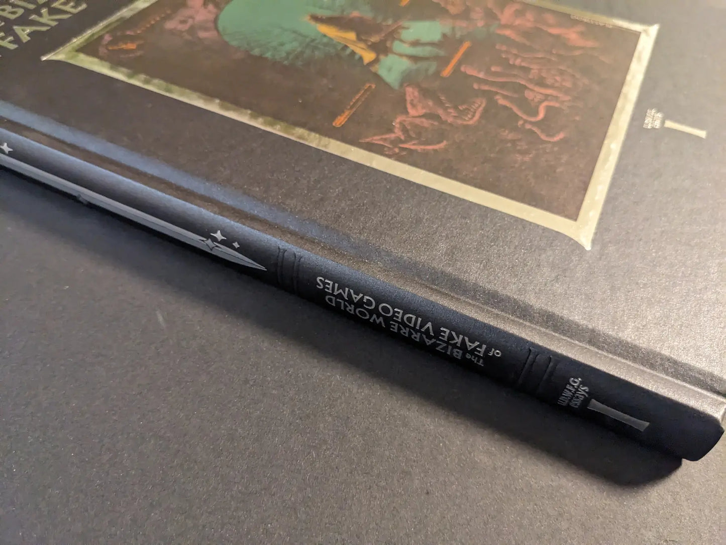 Angled close-up of the spine of The Bizarre World of Fake Video Games hardcover, featuring the title and a silver sword icon on textured black cloth.