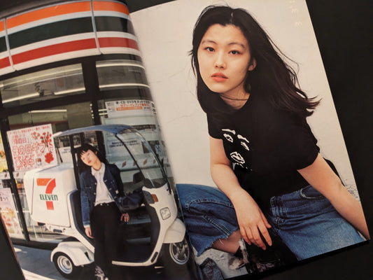 A young woman in a black T-shirt sitting, with a 7-Eleven scooter and storefront in the background.