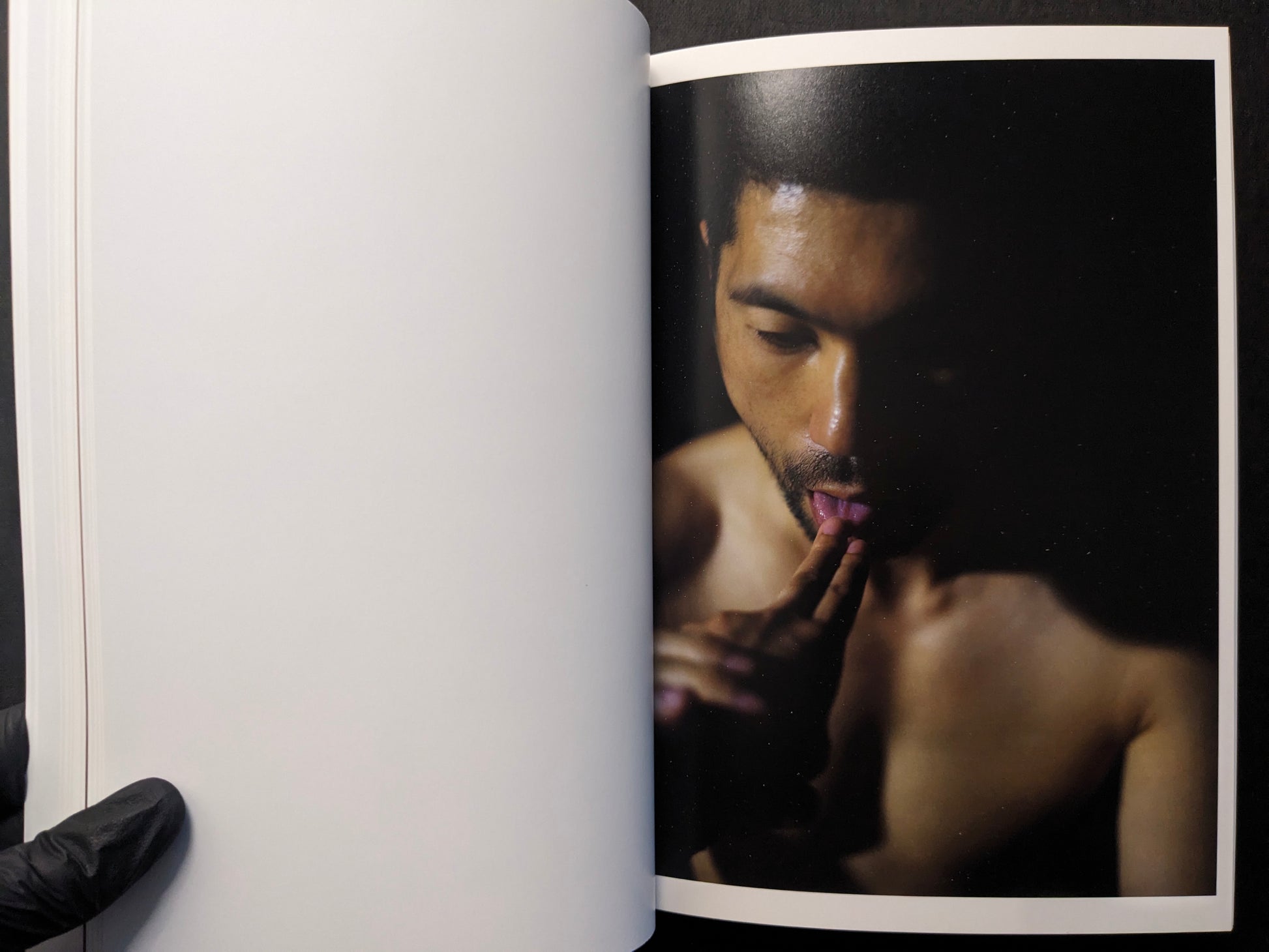 Interior photograph of a shirtless man in low light, hand near mouth.