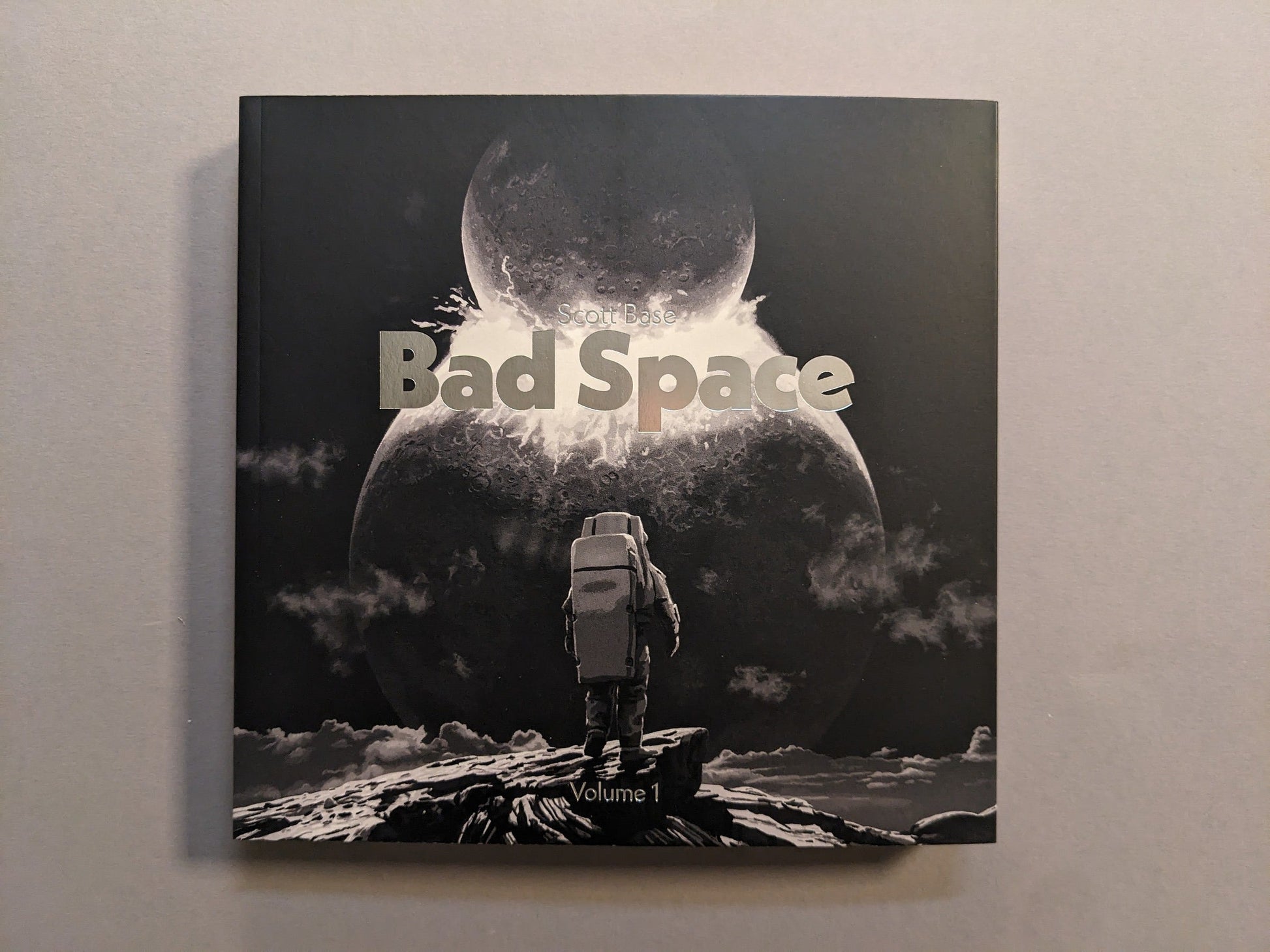 Front cover of Bad Space – Volume 1 by Scott Base with hot silver stamping; astronaut facing a split moon; softcover, 20×20 cm.
