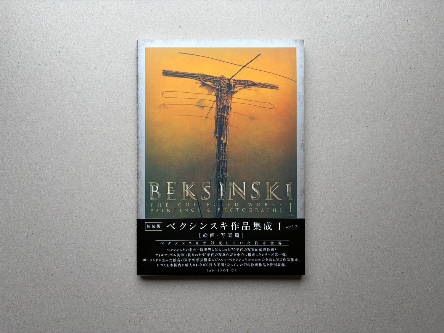 Beksiński - The Collected Works I ver. 1.2 front cover on a grey surface, with an orange painting and the title printed across the lower half.