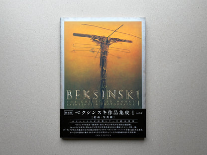 Beksiński - The Collected Works I ver. 1.2 front cover on a grey surface, with an orange painting and the title printed across the lower half.