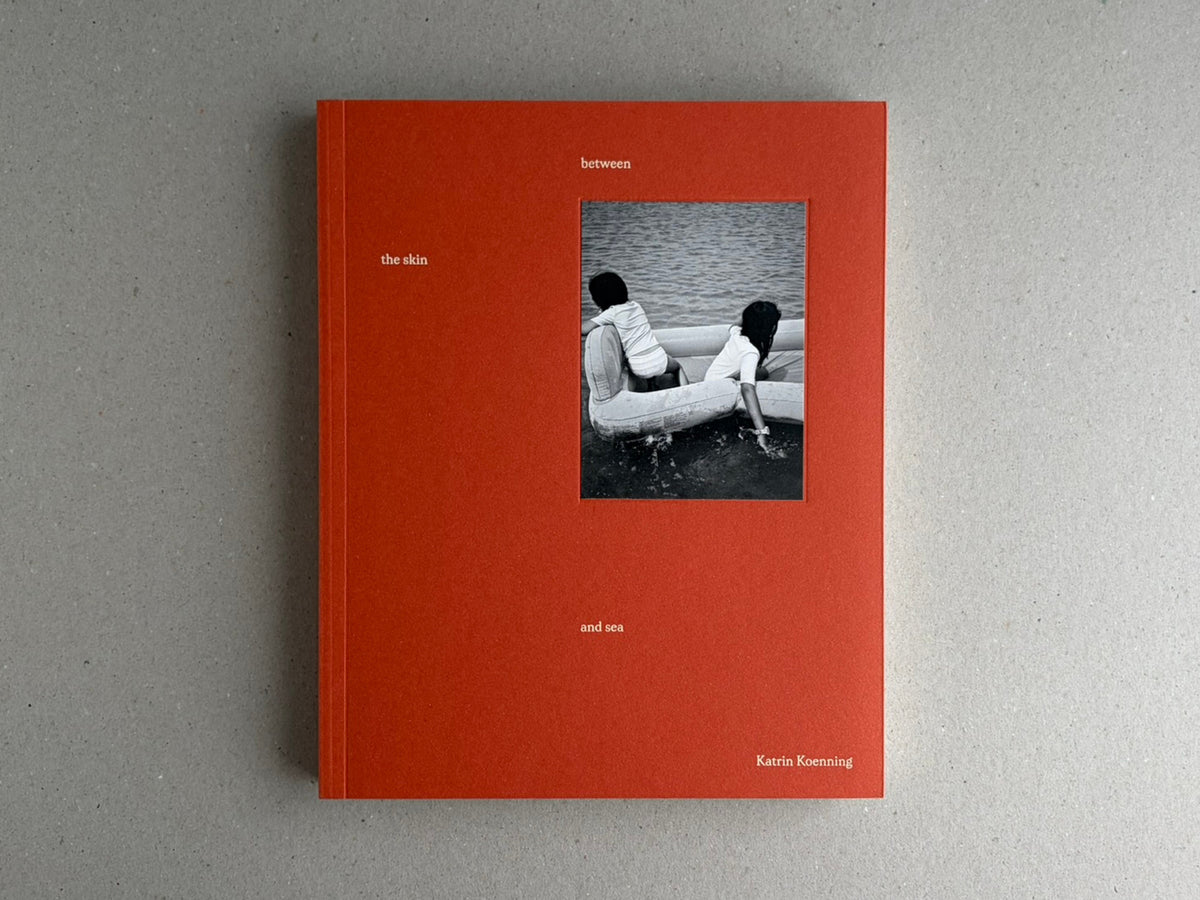 Front cover of between the skin and sea by Katrin Koenning, showing a red-orange cover with a small black-and-white photograph of two children in a boat on water.