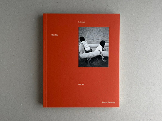 Front cover of between the skin and sea by Katrin Koenning, showing a red-orange cover with a small black-and-white photograph of two children in a boat on water.