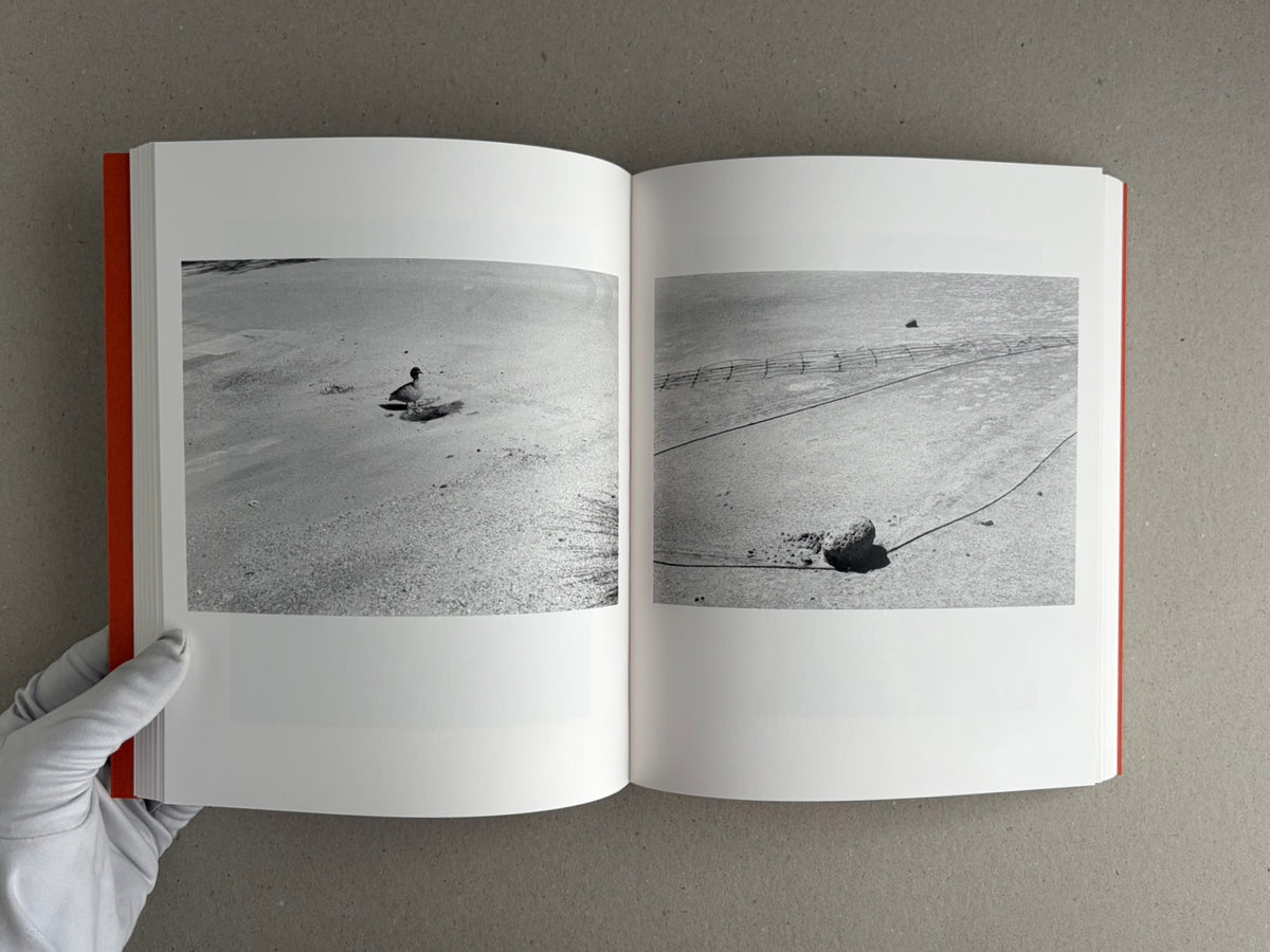 Open spread of between the skin and sea by Katrin Koenning, showing two black-and-white photographs of a shoreline with small objects and lines in the sand.