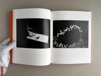 Open spread from between the skin and sea by Katrin Koenning, showing two black-and-white photographs on facing pages: a foot on a ledge at left and a curved branch of leaves at right.