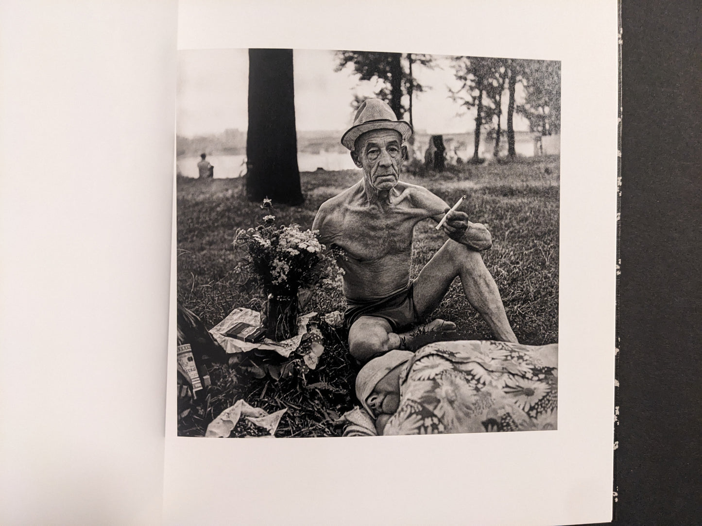 Interior photo from Cheryomushki: older man seated on grass with hat and cigarette, lakeside setting in background.