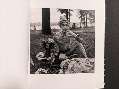 Interior photo from Cheryomushki: older man seated on grass with hat and cigarette, lakeside setting in background.