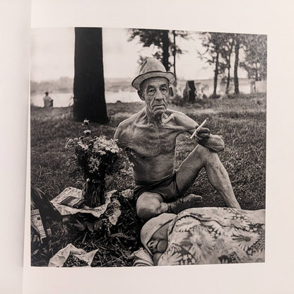 Interior photo from Cheryomushki: older man seated on grass with hat and cigarette, lakeside setting in background.