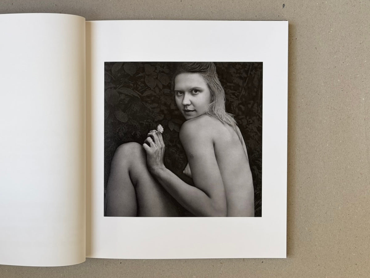 Open book spread showing a black-and-white portrait of a nude woman seated against foliage, holding a small flower.