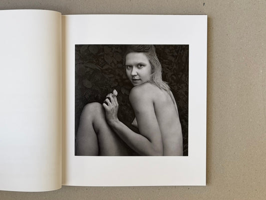 Open book spread showing a black-and-white portrait of a nude woman seated against foliage, holding a small flower.