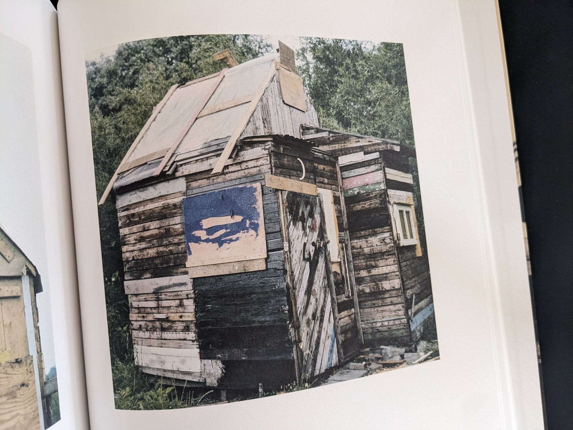Black-and-white photograph of a weathered wooden hut with a pitched roof and patched walls.