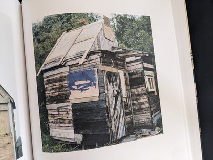 Black-and-white photograph of a weathered wooden hut with a pitched roof and patched walls.