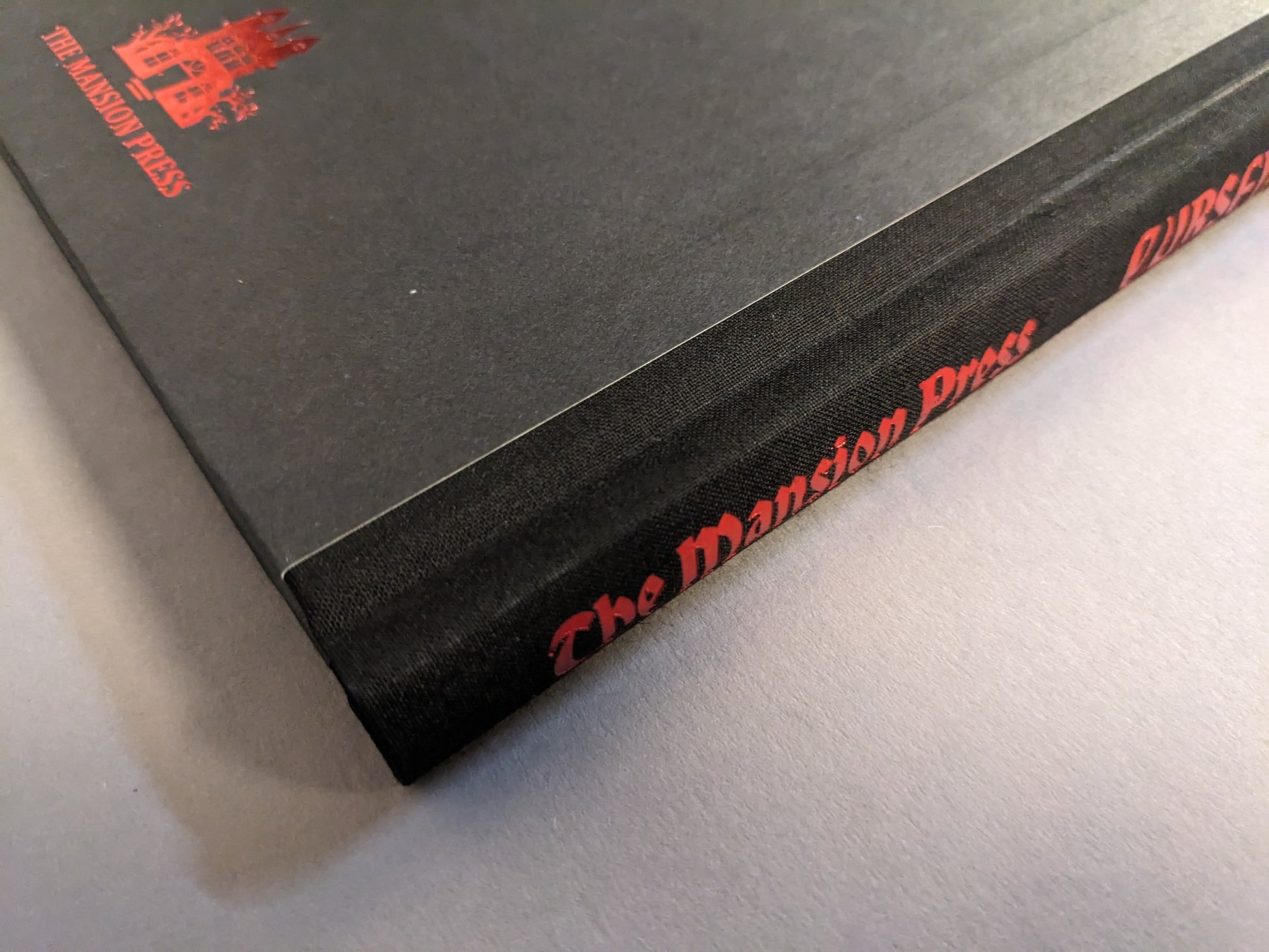 Close-up of the Cursed Swords hardcover cloth spine with red foiled “The Mansion Press” imprint.