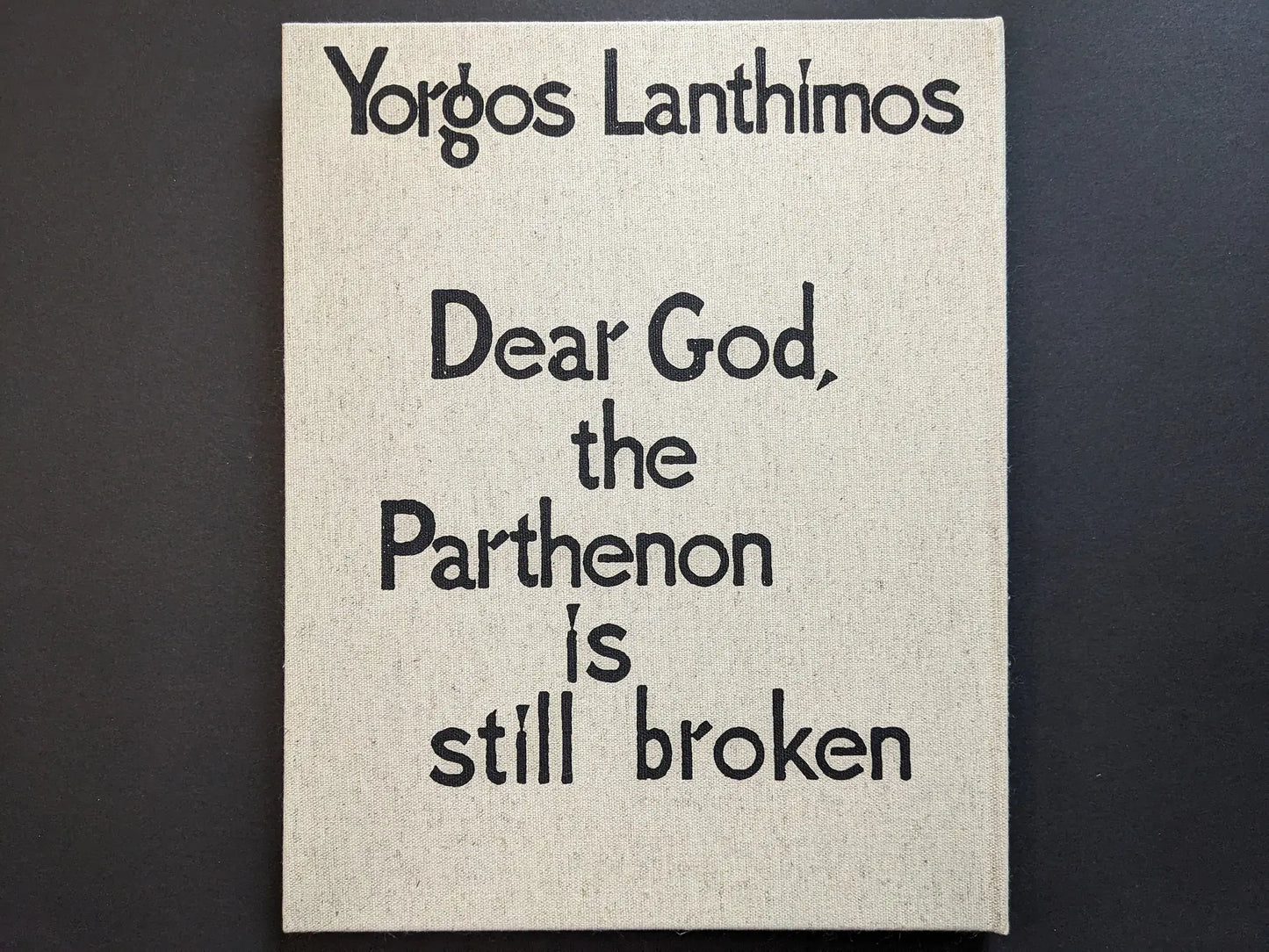 Rear cover of Dear God, the Parthenon is still broken with a black mask motif on natural linen.