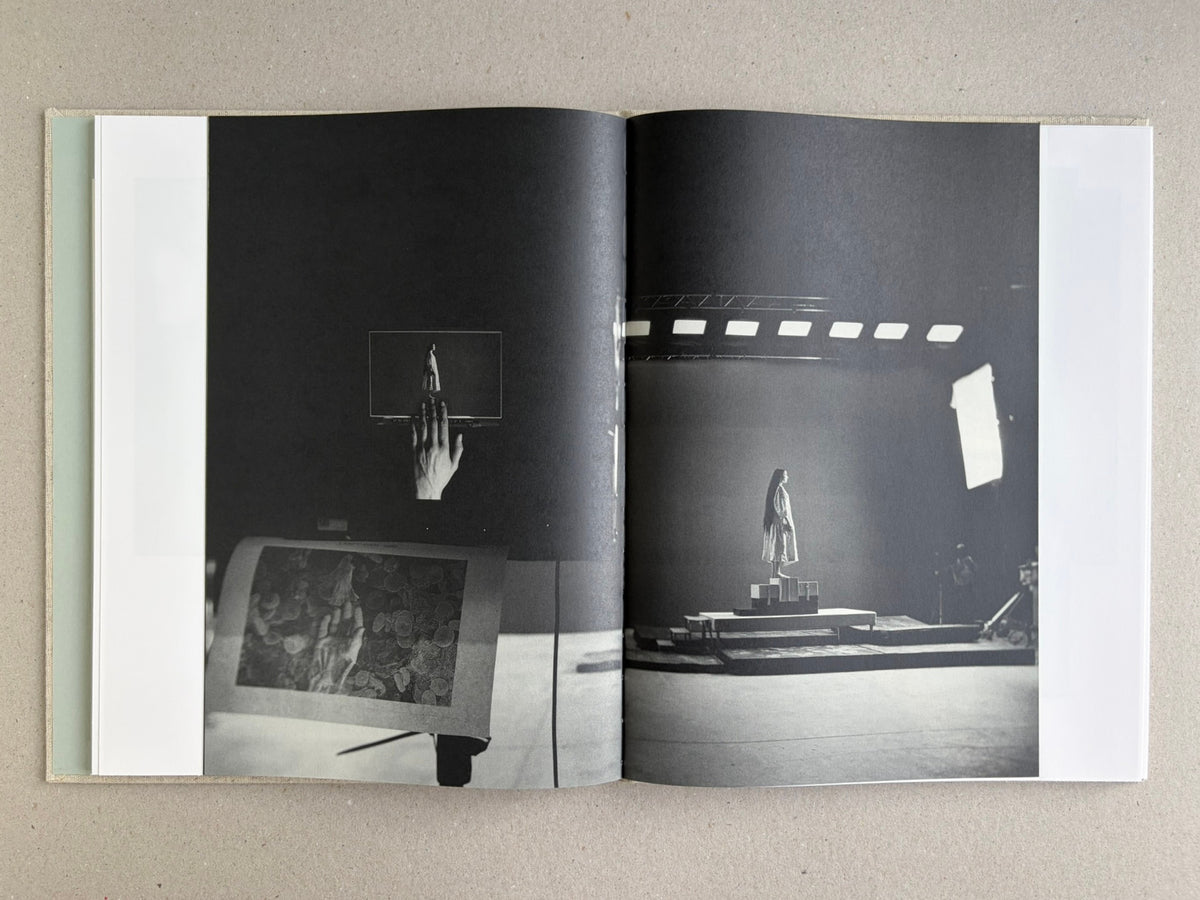 Black-and-white studio spread from Dear God, the Parthenon is still broken, with a hand holding a small figure on the left and a lit stage setup on the right.