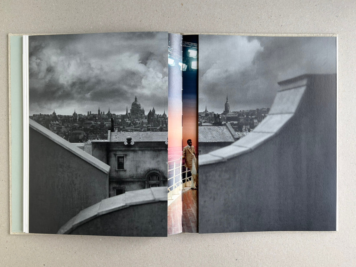 Gatefold spread in Dear God, the Parthenon is still broken, with a black-and-white city view and a narrow central strip showing a figure on a ship deck.