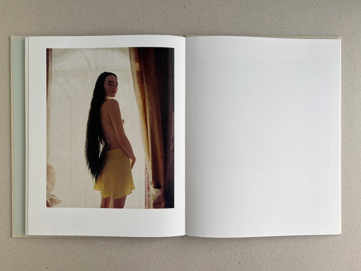 Colour portrait of a standing woman in Dear God, the Parthenon is still broken, framed by curtains and backlight.