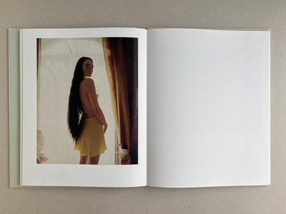 Colour portrait of a standing woman in Dear God, the Parthenon is still broken, framed by curtains and backlight.