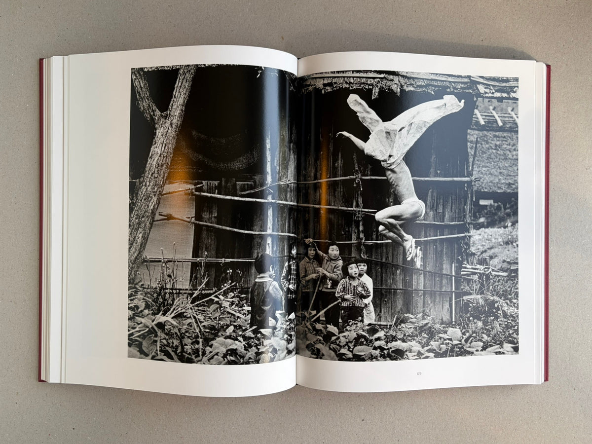 Open spread from Eikoh Hosoe, showing a black-and-white photograph of a figure suspended upside down above several children in front of a wooden building.