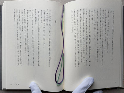 An open book Grass Labyrinth by Takato Yamamoto with Japanese text spread across both pages, held from below by gloved hands. A purple and green ribbon bookmark loops through the center of the pages.