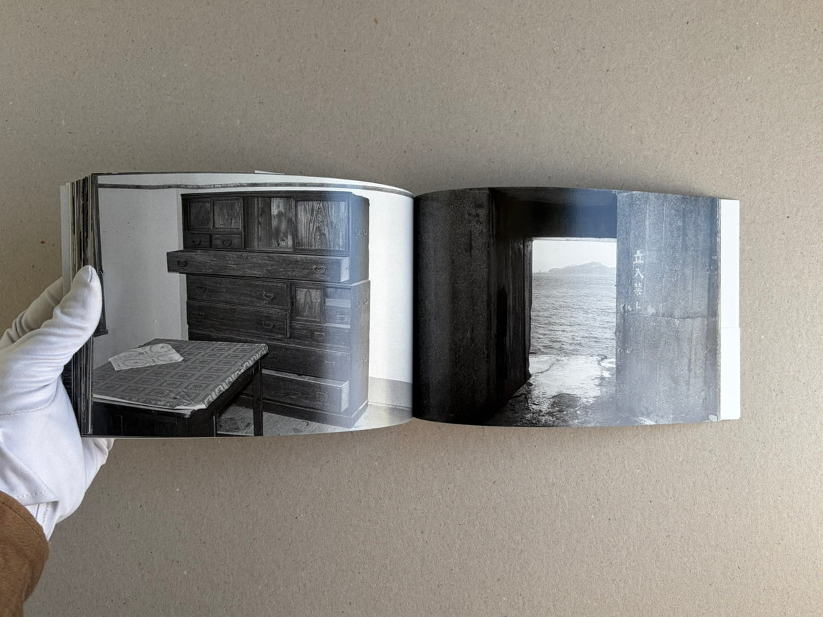 Open copy of Hashima by Taiji Matsue showing a two-page spread: an interior room with a dark wooden chest and table on the left, and a doorway opening to the sea on the right.