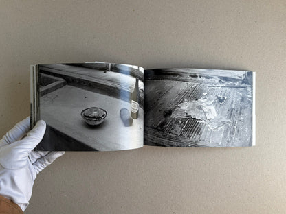 Open spread from Hashima - Taiji Matsue, showing two black-and-white photographs of objects on worn surfaces, held open by a gloved hand.