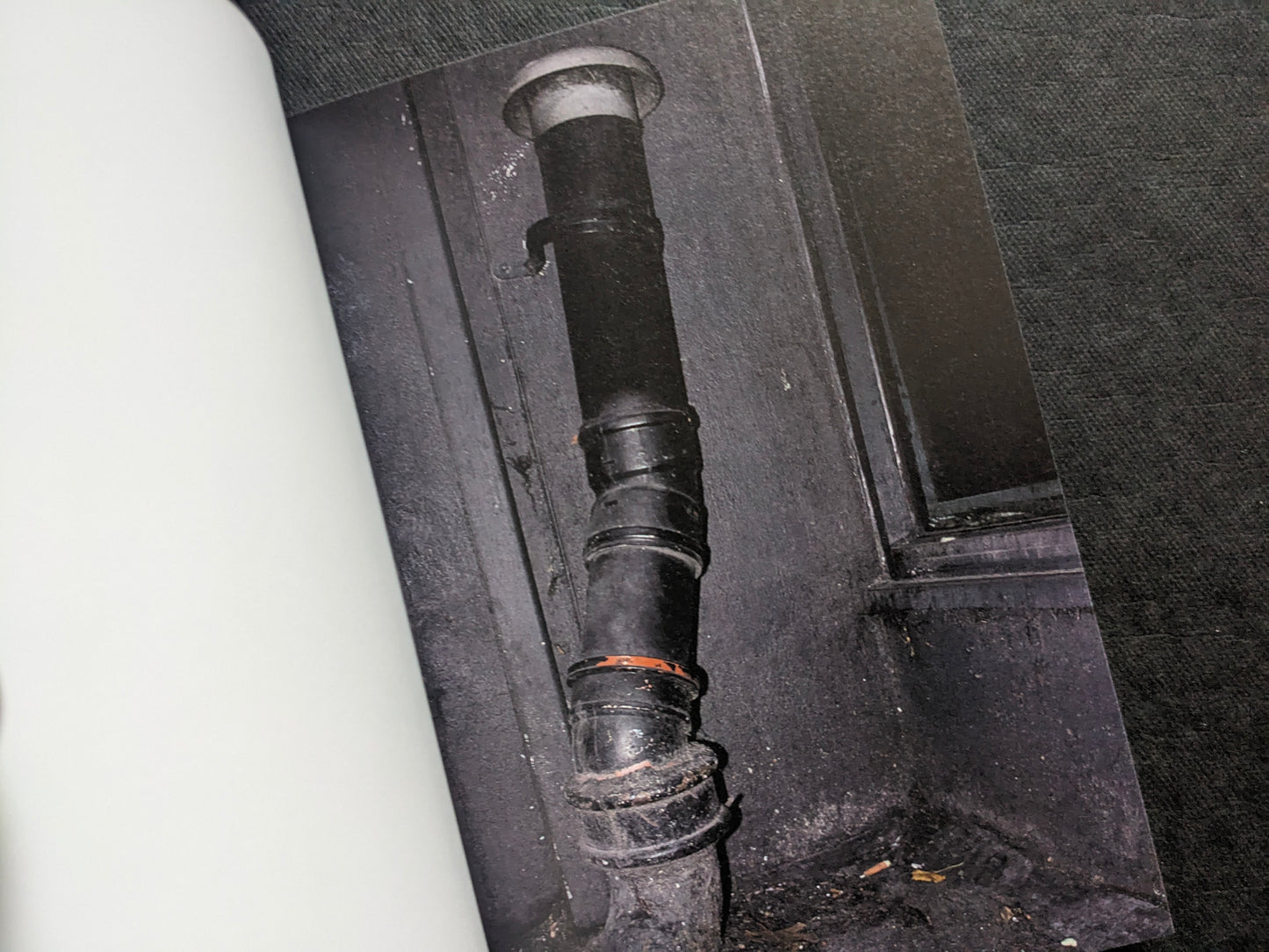 Close-up photograph of a dark drainpipe and wall detail, from I’ll Bet The Devil My Head by Carlos Alba.