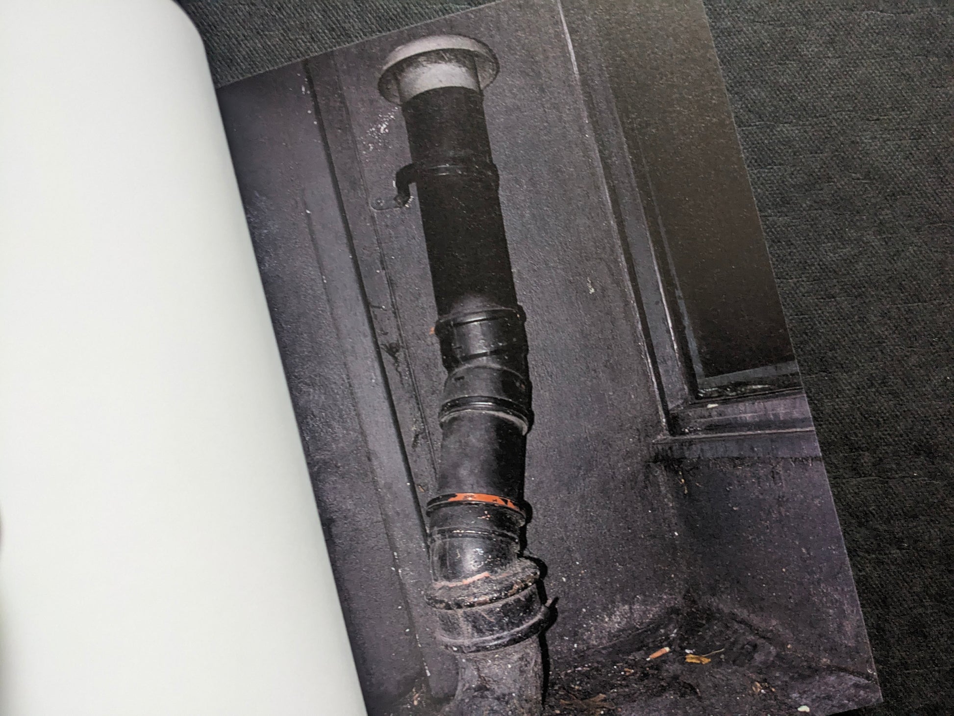 Close-up photograph of a dark drainpipe and wall detail, from I’ll Bet The Devil My Head by Carlos Alba.