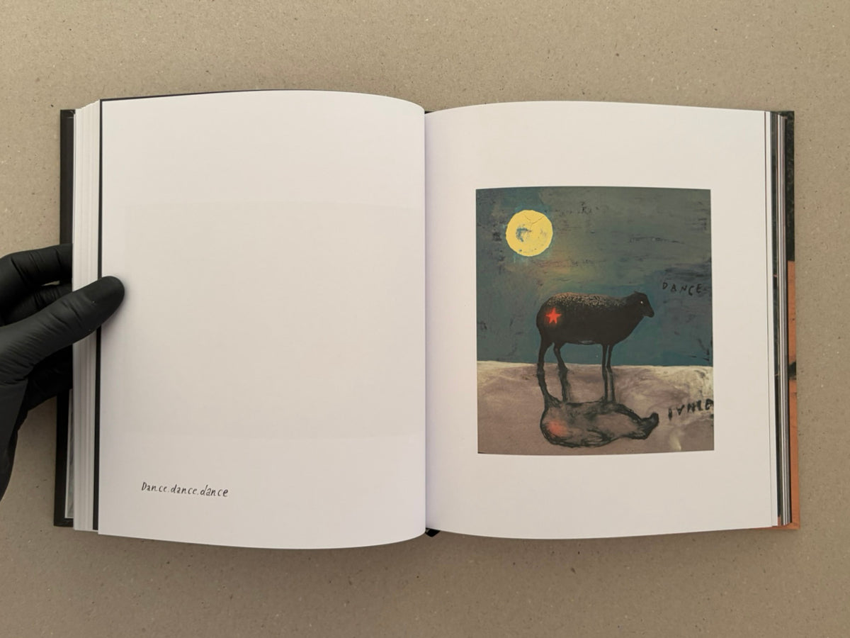 Open artbook spread from In a Dark Time, the Eye Begins to See – The Art of Julia Soboleva, showing a small collage image of a black sheep beneath a yellow moon on the right page and the handwritten text “Dance.dance.dance” on the left page.