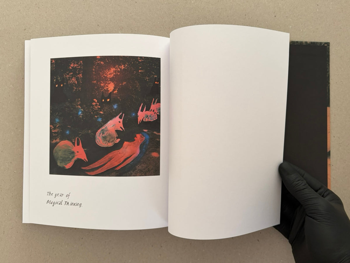 Open artbook spread from In a Dark Time, the Eye Begins to See – The Art of Julia Soboleva, with a dark mixed-media image on the left page and a blank right page; a gloved hand holds the book open.