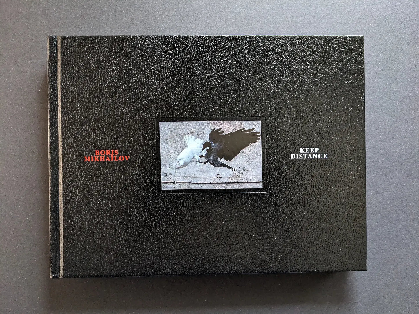 Front cover of Keep Distance by Boris Mikhailov with central photograph of a white bird and a black bird against a black leather-like background.