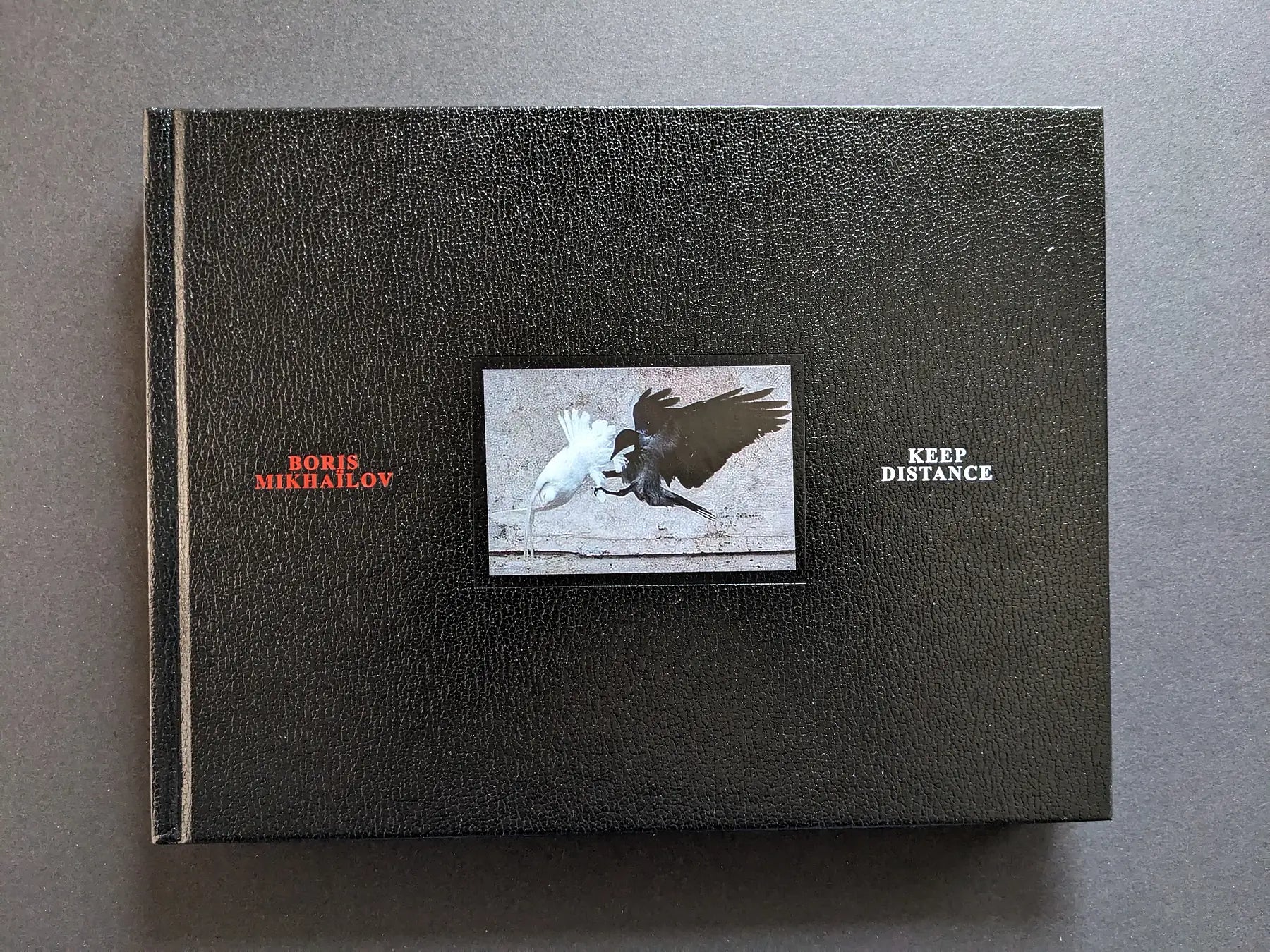 Front cover of Keep Distance by Boris Mikhailov with central photograph of a white bird and a black bird against a black leather-like background.