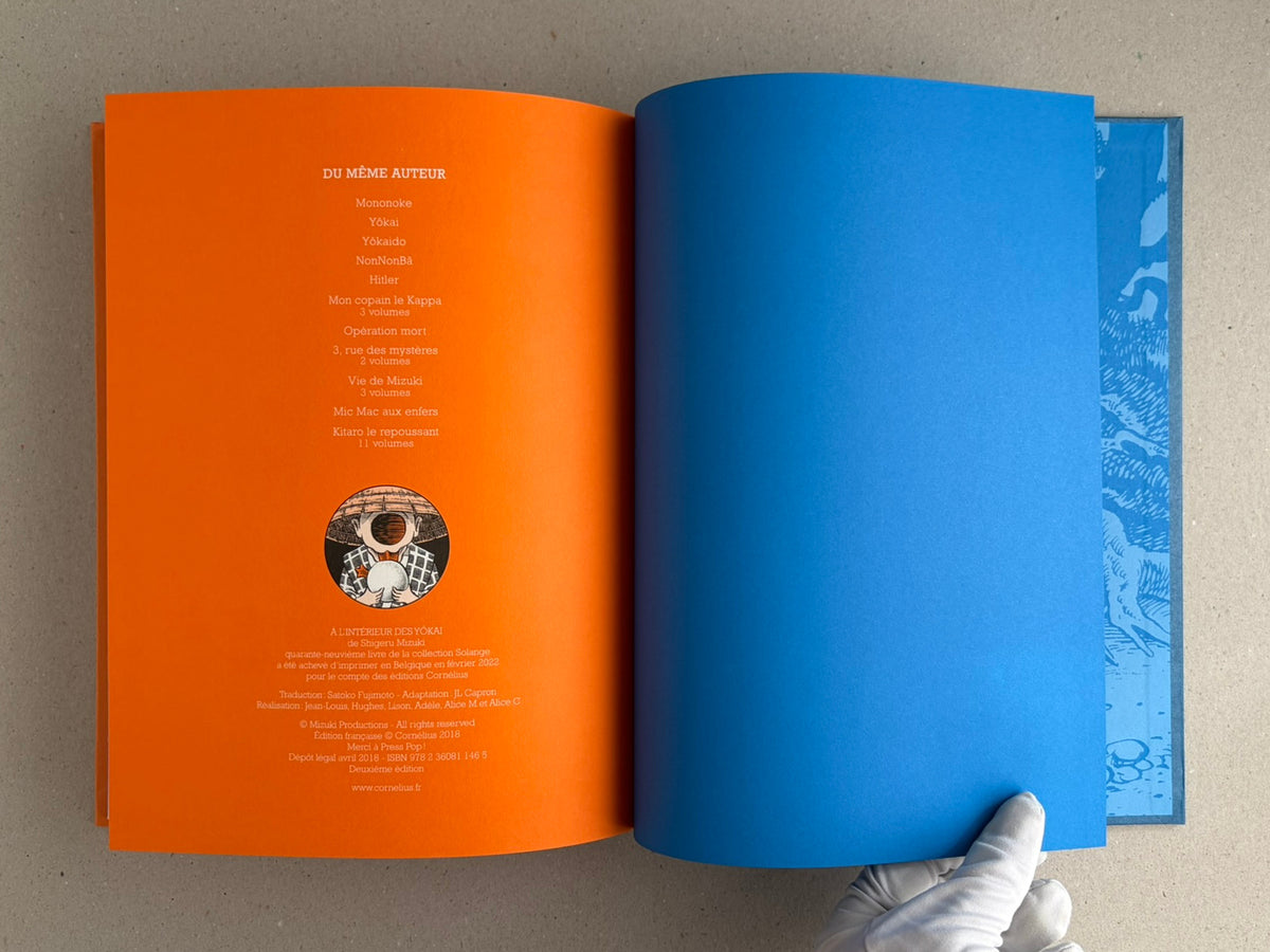 Open book spread from A l'interieur des yokai by Shigeru Mizuki, showing an orange left page with French text and a blue right page, with a gloved hand holding the lower corner.