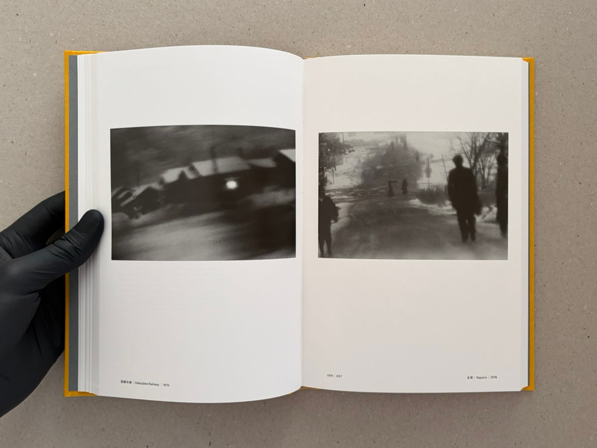 Open spread of Masahisa Fukase 1961–1991 Retrospective showing two black-and-white photographs on white pages, with a gloved hand holding the book open.