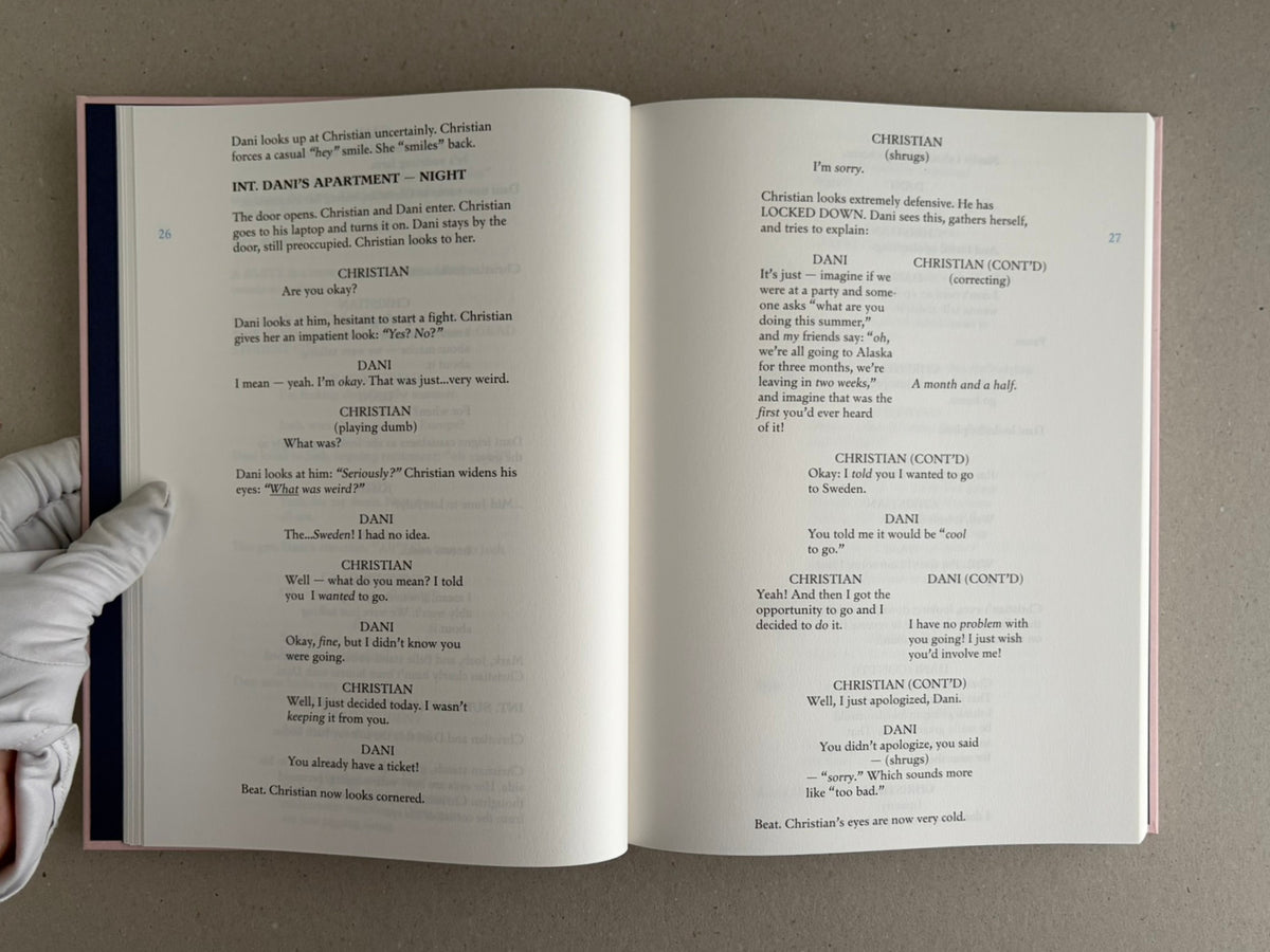 Open spread of the Midsommar screenplay book showing pages 26–27, with screenplay dialogue and a gloved hand holding the lower left corner.