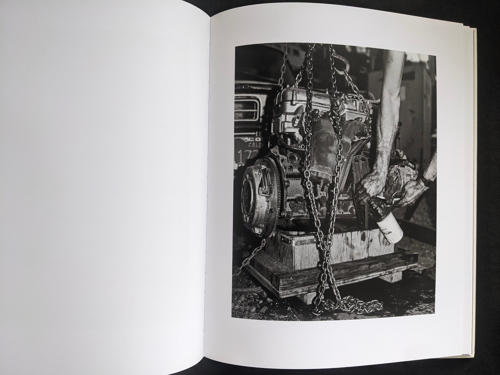 Interior photograph from Mimi Plumb’s Landfall showing a mechanic’s hands working on an engine suspended by chains.