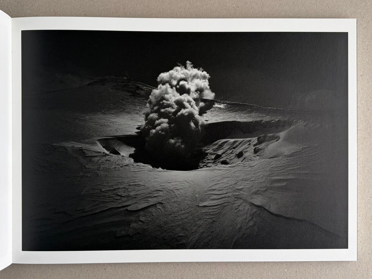 Open book page showing a black-and-white landscape photograph with a central plume of smoke or steam rising from a crater-like depression in snow or sand.