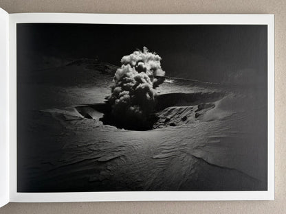 Open book page showing a black-and-white landscape photograph with a central plume of smoke or steam rising from a crater-like depression in snow or sand.