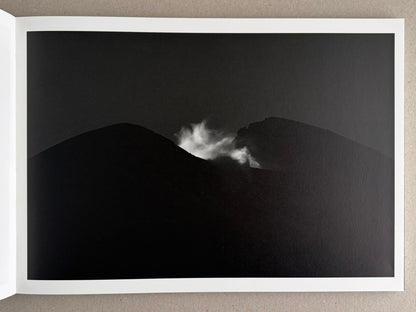 Open photobook spread from New Land: Pre-Hokkaido Landscapes by Toshiki Nakanishi, showing a black-and-white landscape image with dark hills and a plume of steam or mist between them.