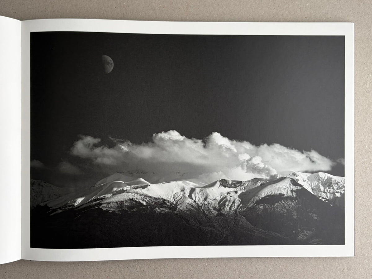 Open book spread showing a black-and-white landscape photograph of snow-covered mountains under a dark sky, with the moon visible above the peaks.