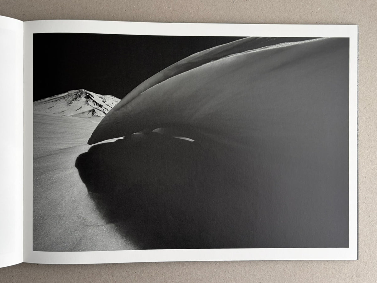 Open photobook spread from New Land: Pre-Hokkaido Landscapes by Toshiki Nakanishi, showing a black-and-white landscape photograph with a snow-covered mountain in the distance and a large dark curved form in the foreground.