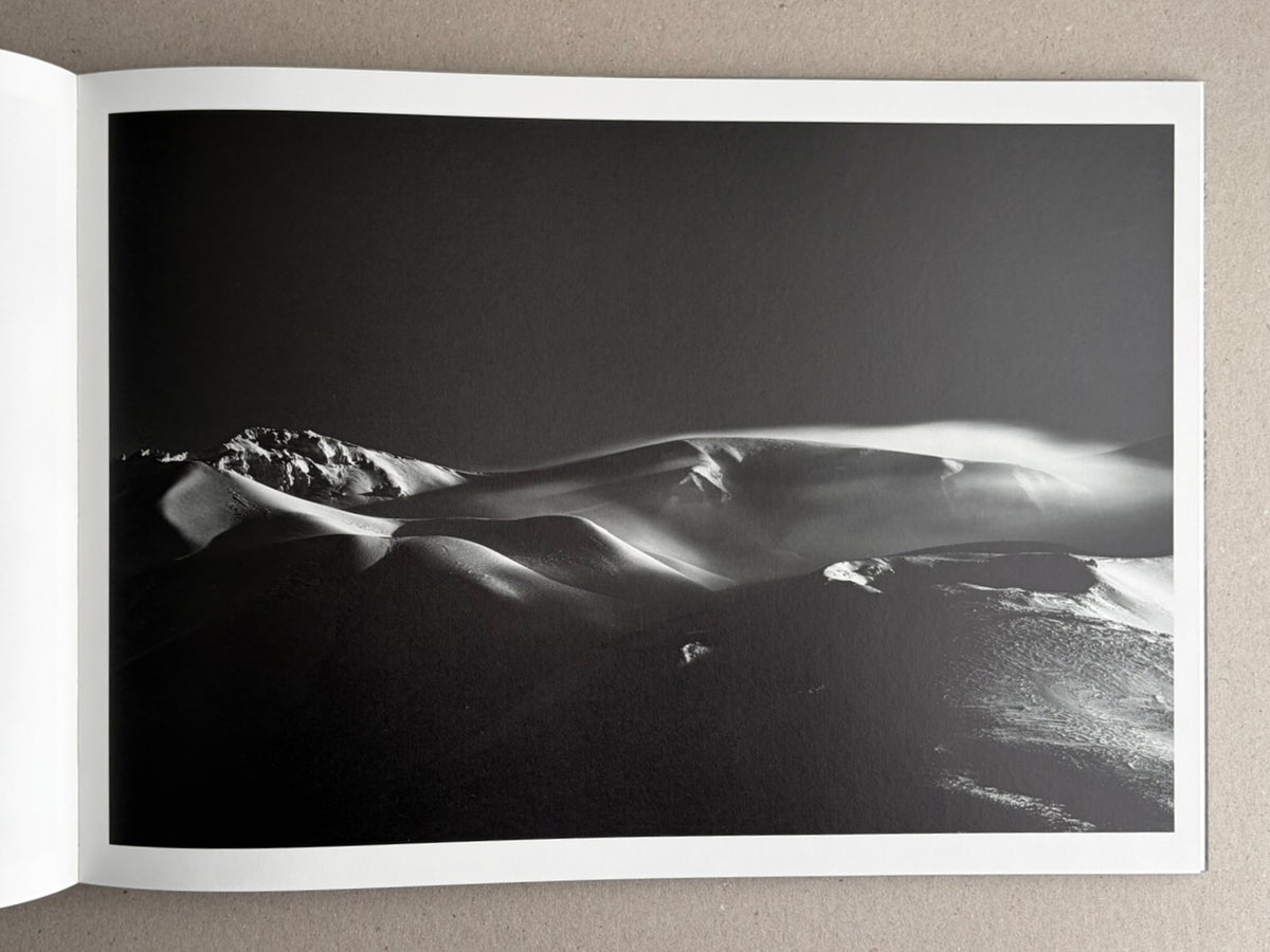 Open photobook spread showing a black-and-white landscape photograph of snow-covered ridges and drifting light.