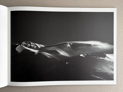 Open photobook spread showing a black-and-white landscape photograph of snow-covered ridges and drifting light.