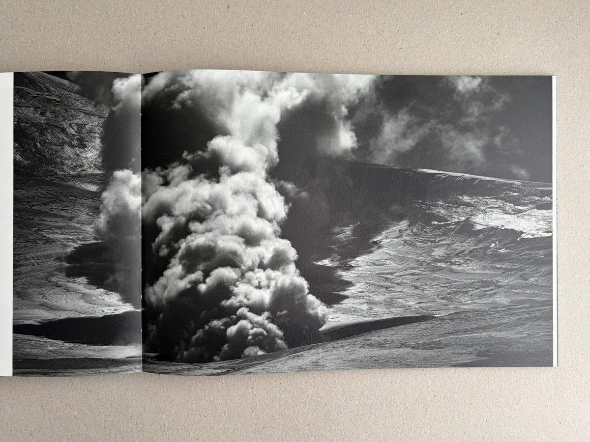 Open photobook spread from New Land: Pre-Hokkaido Landscapes by Toshiki Nakanishi, showing a black-and-white landscape image with a dense plume of smoke or steam rising between dark slopes.