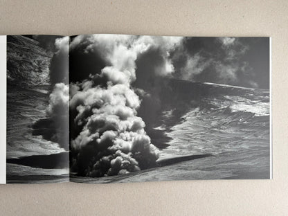 Open photobook spread from New Land: Pre-Hokkaido Landscapes by Toshiki Nakanishi, showing a black-and-white landscape image with a dense plume of smoke or steam rising between dark slopes.