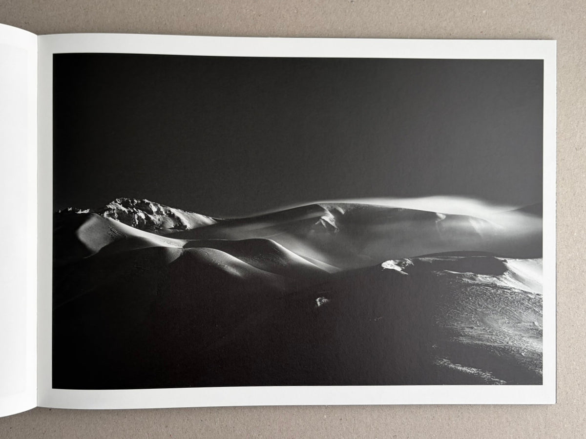 Open photobook spread showing a black-and-white landscape photograph of snow-covered dunes or ridges under a dark sky.